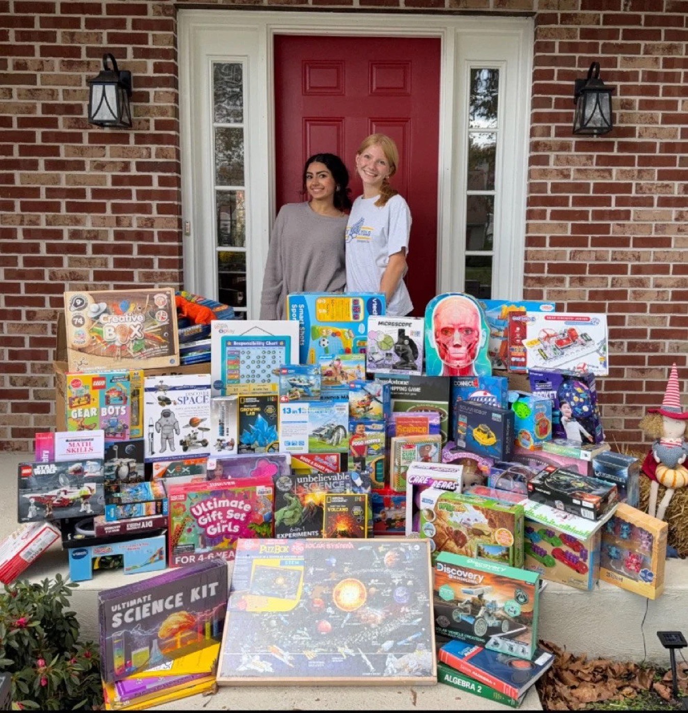 Two STEM Spark volunteers standing on a porch beside a large collection of donated STEM toys and science kits.