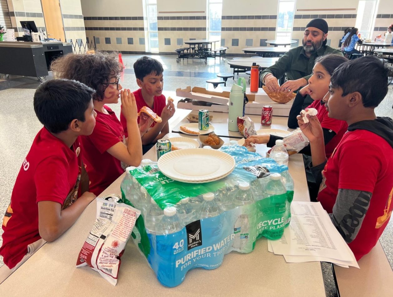 A mentor sitting with a group of students around a lunchroom table during a STEM Spark session.